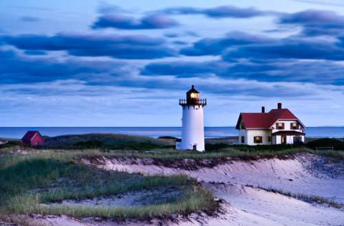 A lighthouse stands beside a house on sandy dunes, with a calm ocean and cloudy sky in the background at dusk.