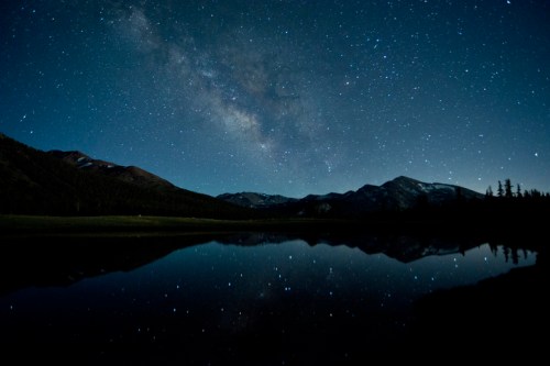A serene night sky filled with stars and the Milky Way, reflecting on a calm lake surrounded by mountains.