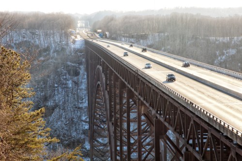 A bridge spans a valley with cars driving on it, surrounded by trees and a snowy landscape in the background.