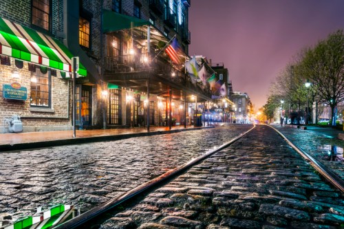 A cobblestone street at night, lined with buildings and reflecting lights, creating a vibrant urban atmosphere.