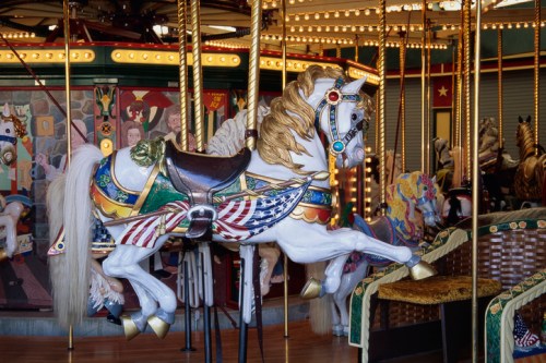 A beautifully painted carousel horse with a colorful saddle, surrounded by other carousel horses in a vibrant setting.