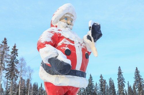 A large, snow-covered Santa Claus statue holding a black object, set against a blue sky and pine trees.