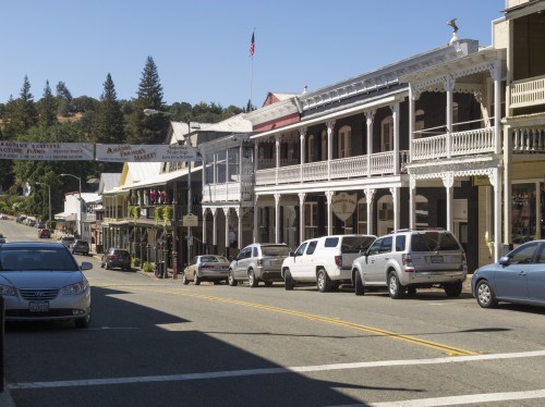 Historic buildings line a street in a small town, with parked cars and trees in the background under a clear blue sky.