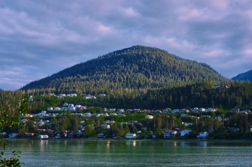 A serene lake reflects a forested mountain, with a small town nestled along the water's edge under a cloudy sky.