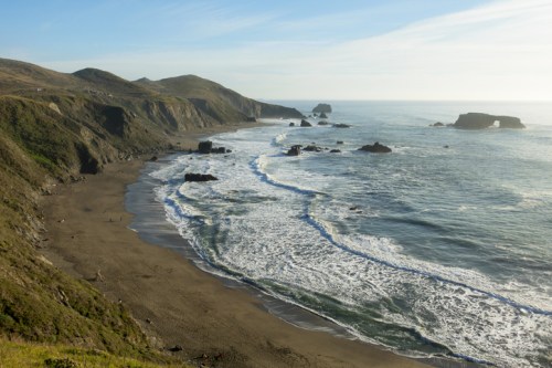 A serene coastal view with waves gently lapping a sandy beach, surrounded by rolling hills and rocky outcrops.