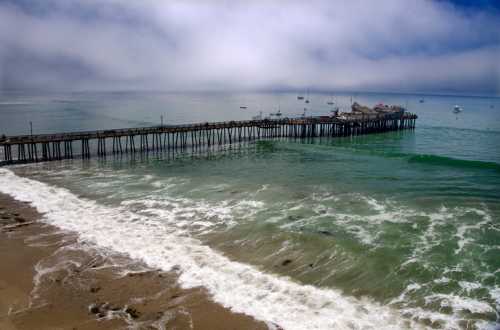 A wooden pier extends into a calm ocean under a cloudy sky, with gentle waves lapping at the sandy shore.