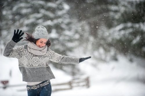 A woman in a cozy sweater and hat joyfully dances in the snow, surrounded by a winter landscape.