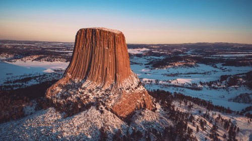 Aerial view of a snow-covered butte with steep, layered sides, surrounded by a winter landscape.