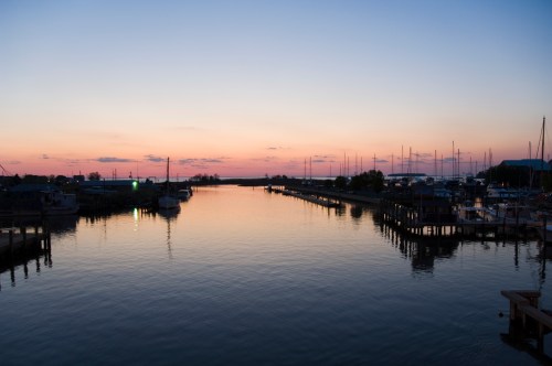 A serene sunset over a calm waterway, with boats docked along the shore and a colorful sky reflecting on the water.