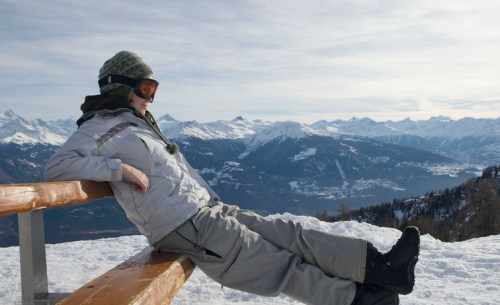 A person in winter gear relaxes on a bench, overlooking a snowy mountain landscape under a clear sky.