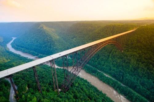 Aerial view of a tall bridge spanning a river, surrounded by lush green mountains under a colorful sunset.