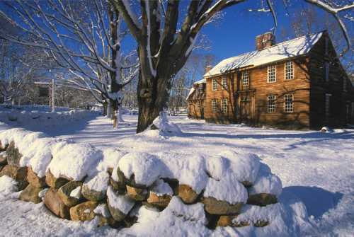 A snow-covered landscape featuring a wooden house and a stone wall, surrounded by bare trees under a clear blue sky.