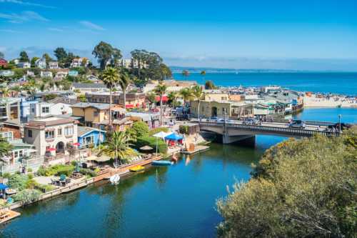 A scenic view of a coastal town with colorful buildings, palm trees, and a bridge over a calm waterway leading to the beach.