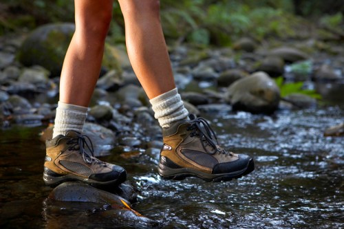 A person stands on rocks in a stream, wearing hiking boots and white socks, with water flowing around their feet.