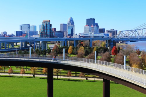 A scenic view of a city skyline with a bridge, park, and colorful trees under a clear blue sky.