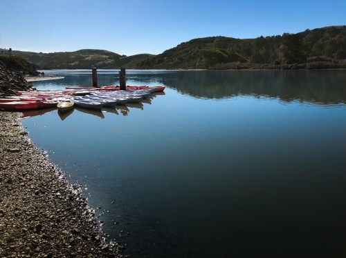 A calm lake with colorful kayaks lined up on the shore, surrounded by green hills under a clear blue sky.