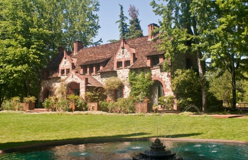A charming stone house surrounded by lush greenery and trees, with a fountain in the foreground.