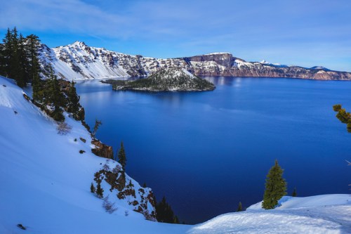 A snowy landscape overlooking a deep blue lake surrounded by mountains and evergreen trees.