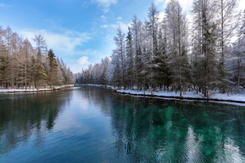 A serene river surrounded by snow-covered trees under a blue sky, reflecting the tranquil landscape.