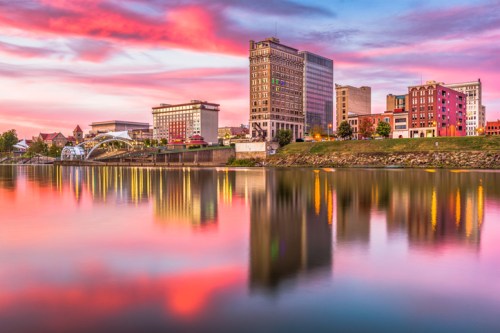 City skyline at sunset, reflecting in the water with colorful clouds and illuminated buildings.
