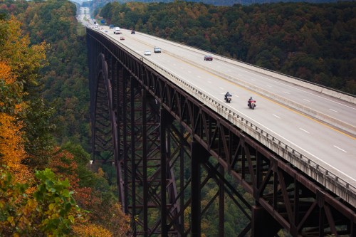 A high bridge spans a valley, with cars and motorcycles traveling along it, surrounded by autumn-colored trees.