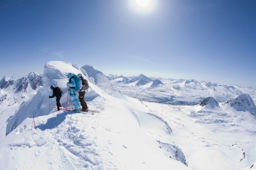 Two climbers on a snowy mountain peak, surrounded by a vast landscape of snow-covered mountains under a clear blue sky.