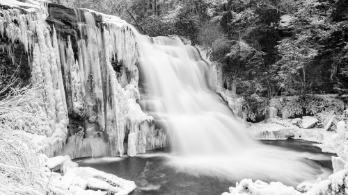 A frozen waterfall surrounded by snow-covered trees, captured in a serene black-and-white scene.