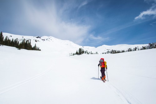 A skier with a backpack ascends a snowy landscape under a blue sky, surrounded by mountains and trees.