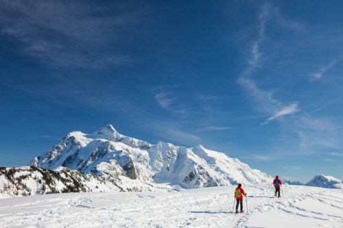 Two skiers traverse a snowy landscape with majestic mountains under a clear blue sky.