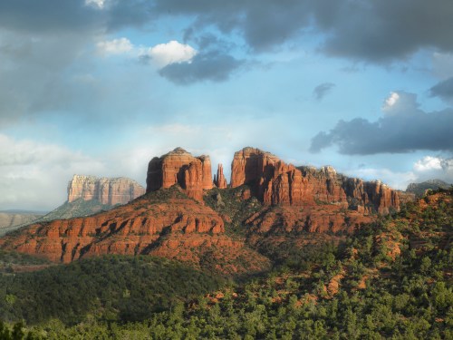 Red rock formations under a cloudy sky, with lush greenery in the foreground and distant mountains in the background.