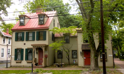 A two-story house with a red metal roof, green shutters, and a porch, surrounded by trees and power lines.