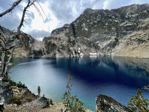 A serene mountain lake surrounded by rocky cliffs and trees, reflecting a cloudy sky.