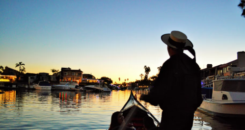 A gondolier rows a boat at sunset, with houses and palm trees reflected in the calm water.