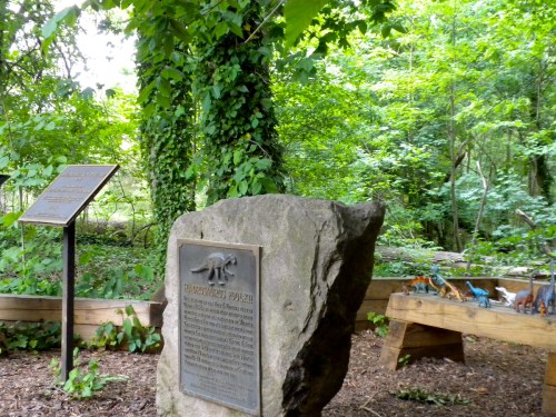 A stone plaque in a wooded area, surrounded by trees and a display of dinosaur figures on a wooden bench.