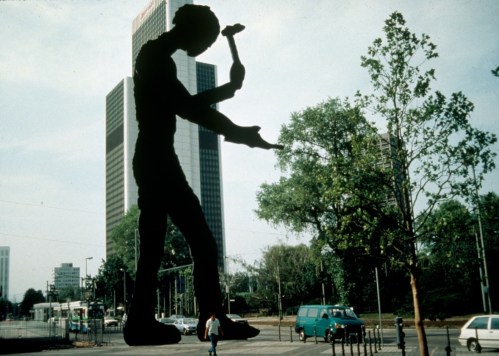 A large silhouette of a worker holding a hammer stands in front of modern buildings and trees in an urban setting.