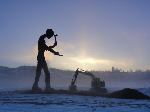 A large silhouette of a man with a hammer stands beside an excavator against a misty, sunset backdrop.