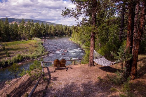A serene riverside scene with a hammock between trees and a wooden chair overlooking the flowing water and lush greenery.