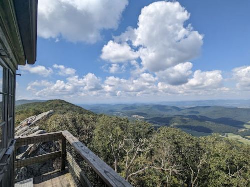A scenic view from a mountain lookout, featuring rolling hills and a blue sky with fluffy clouds.