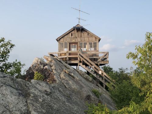 A wooden lookout tower on a rocky outcrop, surrounded by greenery under a clear sky.
