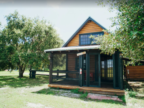 A charming wooden cabin with a porch, surrounded by greenery and trees on a sunny day.
