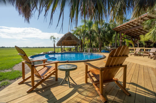 A sunny outdoor pool area with wooden chairs, a table, and palm trees, surrounded by a lush green landscape.