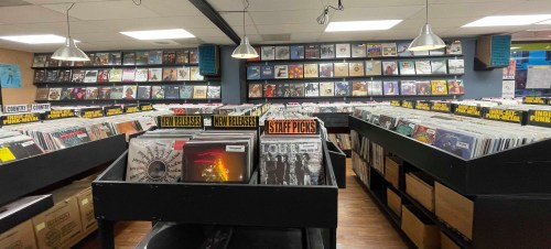 A record store interior with rows of vinyl records, featuring new releases and staff picks prominently displayed.