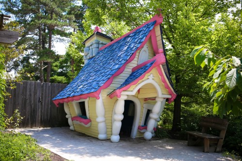 A whimsical, colorful house with a slanted roof, surrounded by greenery and a path leading to a nearby bench.