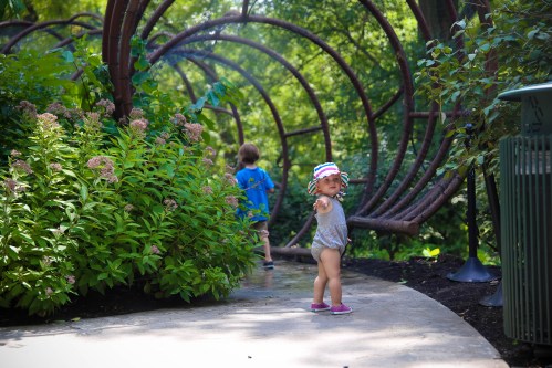 A toddler in a sun hat stands on a path, pointing, while another child walks ahead in a lush garden setting.