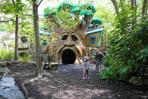 A child explores a whimsical treehouse play structure surrounded by greenery and nature.
