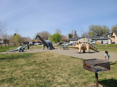 A park scene featuring large dinosaur sculptures, with a child playing nearby and a grill in the foreground.