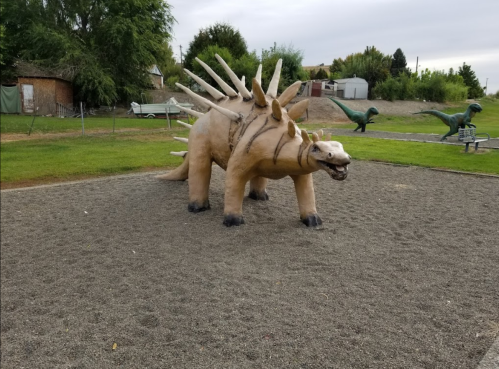 A large dinosaur sculpture resembling a stegosaurus in a playground, surrounded by grass and other dinosaur figures.
