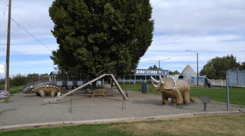 A dinosaur-themed playground featuring dinosaur sculptures, a slide, and a sign for a dino store under a cloudy sky.