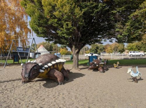 Playground featuring large dinosaur sculptures and a swing set under a tree, with a sunny sky in the background.