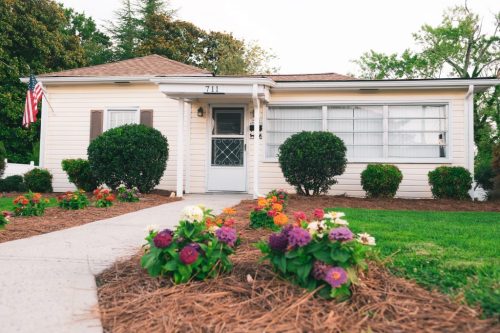 A charming yellow house with a front porch, surrounded by colorful flower beds and a small lawn, featuring an American flag.